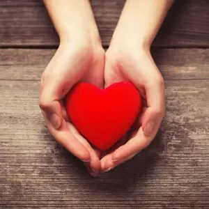Hands gently holding a red heart on a wooden surface.